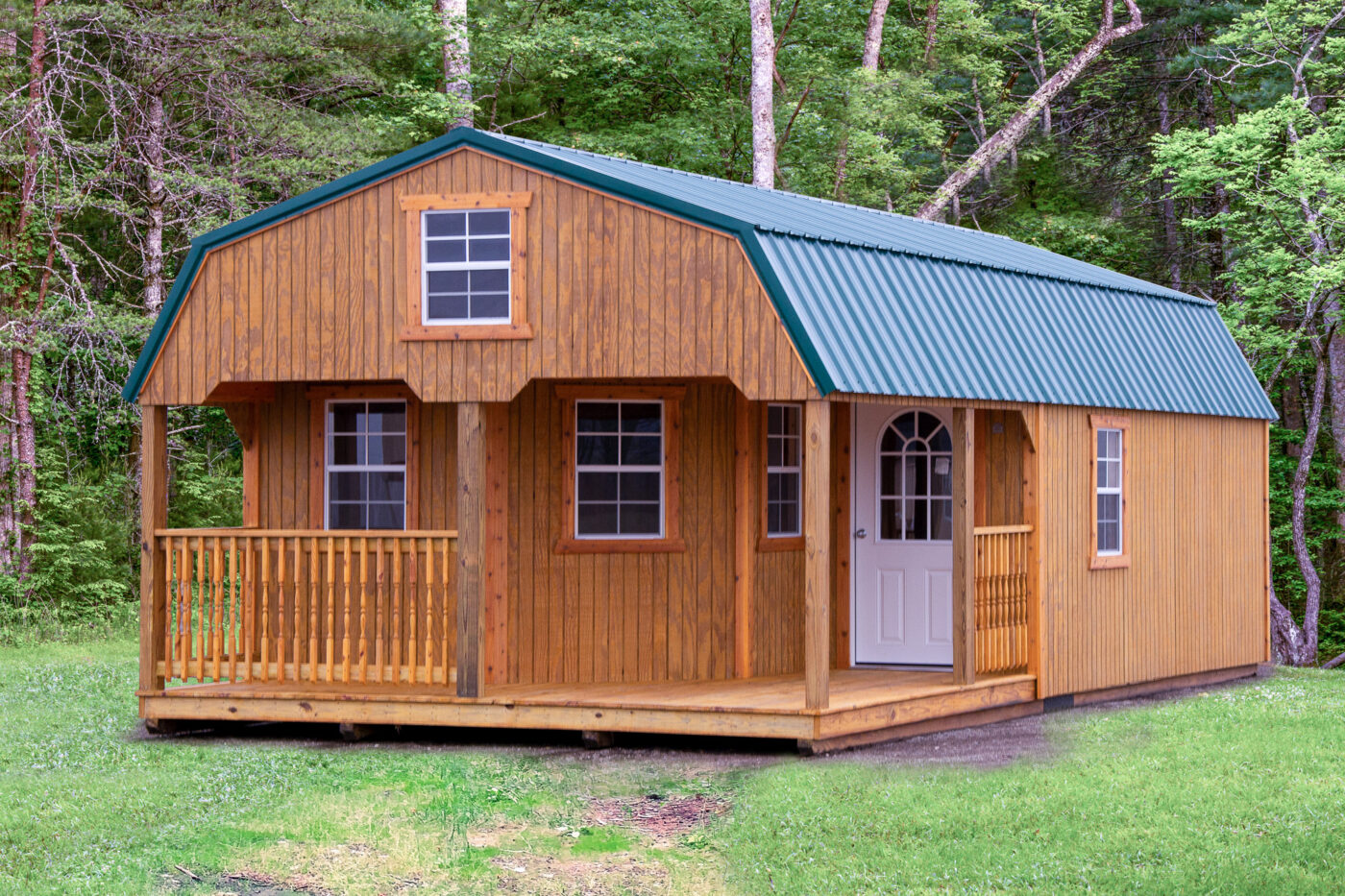 One of our deep golden wood Bayfront lofted sheds for sale in Lewiston MI - Cozy wooden siding with four visible white framed windows and matching white glass-inset door facing partially fenced wood porch beneath green roof - forest trees in background