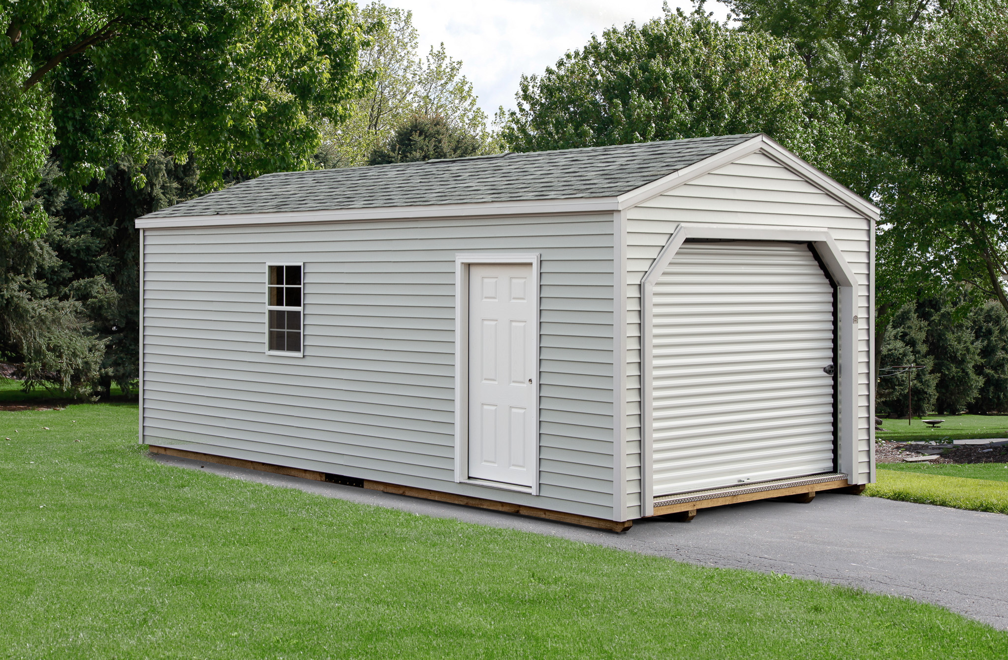 One of our light gray garage sheds for sale in Owosso 2 MI - Gray siding with corrugated overhead door white side door and window beneath gray shingled roof - trees in the background
