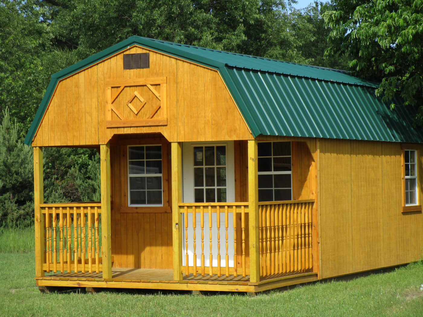 One of our golden wooden lofted cabin sheds for sale in Owosso 2 MI - Golden wood siding and fenced porch white door and three visible windows beneath green metal roof - set on grass beneath luch green trees