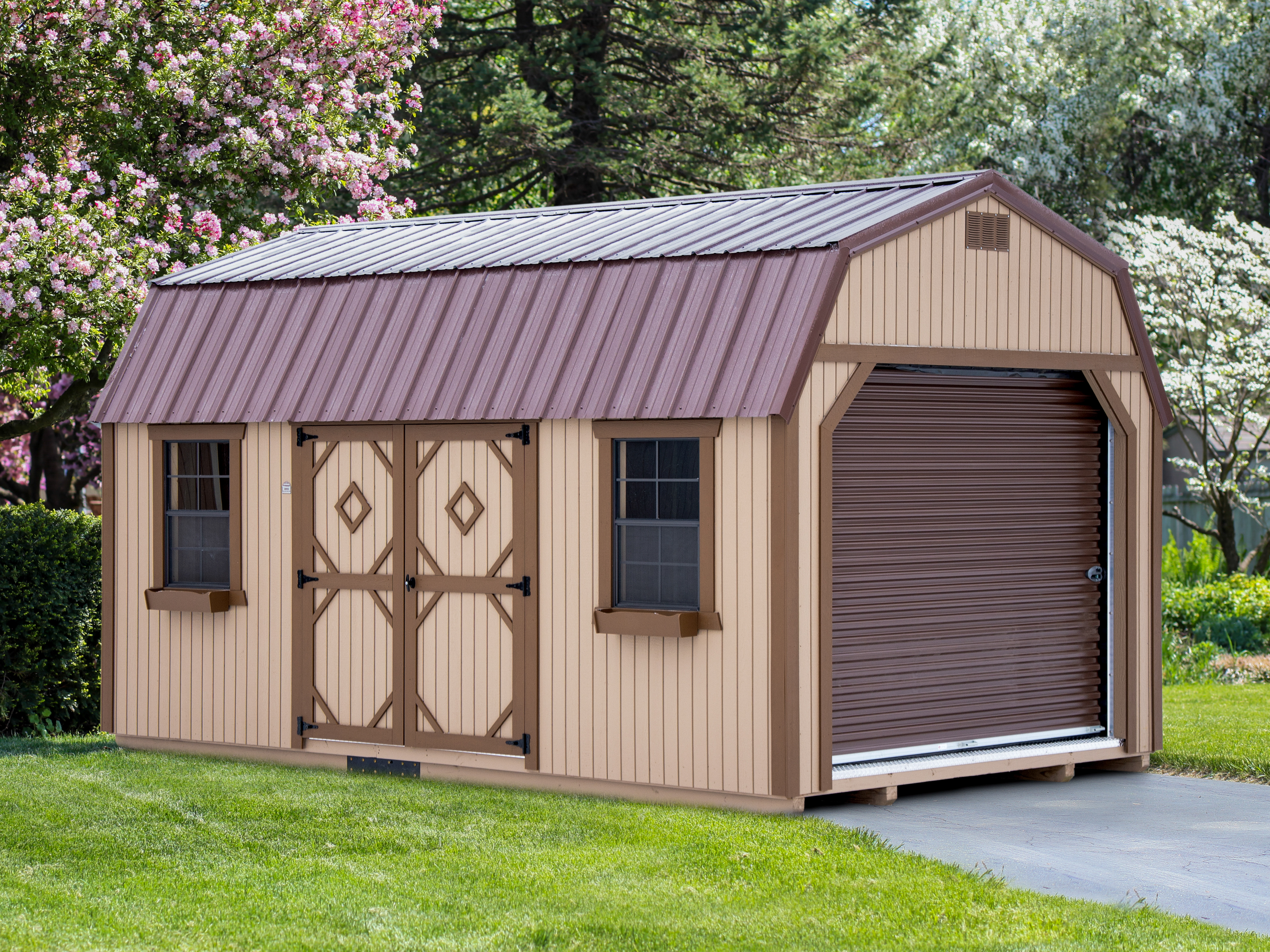 One of our tan and brown lofted portable sheds for sale in Lewiston MI - Tan siding with brown overhead door and brown trimmed double doors and windows under brown roof - shed sits on driveway between areas of grass in front of trees